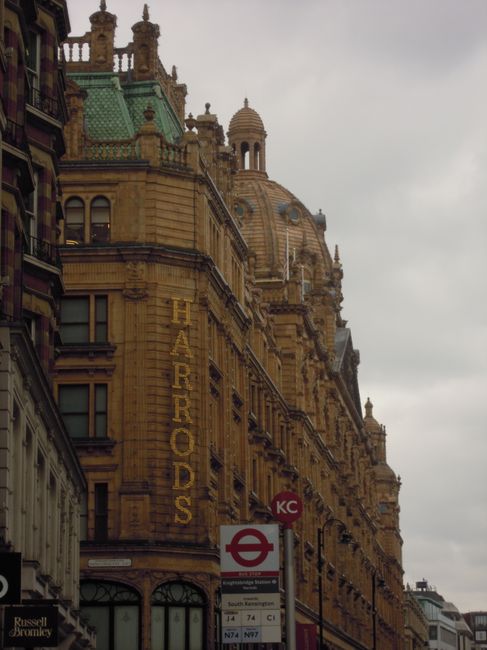 A detailed street scene outside a historic, ornate building with intricate architectural features and topped with a domed roof, located near Bromley North Station. In the foreground, a tall, vertical sign reads 'HARRORDS' in illuminated yellow letters. Below the sign, there is a roundel underground station sign with the red circle and blue bar, indicating the London Underground, alongside a white timetable displaying bus routes, including numbers 74, 74, N74, and N77, posted on a pole. The street scene suggests a busy urban location suitable for occasions involving house removals or furniture transport, with the building potentially serving as a commercial or hotel property. Although no moving activity is directly shown, the proximity to transportation services and the presence of road signage support the context of logistical planning for residential or commercial relocations undertaken by Bromley Man and Van, with the image capturing the environment where lifting, packing, and transportation processes may occur during home relocation or moving services.