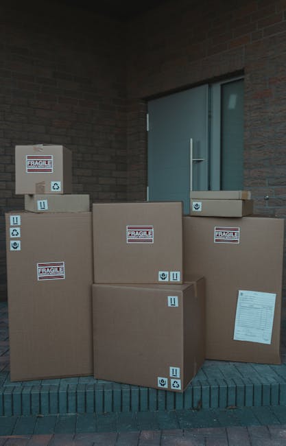 A group of seven large cardboard boxes sealed with packing tape and marked with 'Fragile' stickers and handling symbols, placed on a brick doorstep outside a building with a brick wall and a closed light blue door. The boxes vary in size, with some positioned upright and others lying flat, indicating they are prepared for home relocation or furniture transport. The scene suggests the final stage of packing before loading these items into a van for a house move, with Bromley Man and Van providing the removal service. No people are visible, but the arrangement demonstrates careful packing and readiness for loading during a professional moving process in the context of house removals or packing and moving activities.