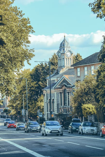 A street scene depicting a church with a prominent clock tower situated behind a row of parked cars, including sedans and hatchbacks, along the pavement. The street is lined with mature trees with green foliage, and lampposts are visible along the roadside. The surrounding buildings include residential houses with brick facades and tiled roofs. The sky is clear with a few scattered clouds, and the area appears to be part of a typical urban neighbourhood in Bromley. This image context aligns with home relocation services, as it shows the environment where a house removal vehicle from Bromley Man and Van may be parked for a same-day moving or furniture transport operation, emphasizing the logistical aspect of house removals in Bromley South.
