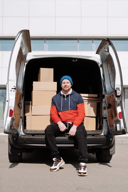 A man wearing a blue beanie, black trainers, dark jeans, and a navy blue and red jacket is sitting on the edge of an open black van, partially loaded with neatly stacked cardboard boxes of various sizes for a home relocation or furniture transport. The van is parked outdoors on a paved surface in front of a modern building with a tiled facade. Inside the van, the back area is visible, showing more boxes and a large cardboard box secured with plastic wrap. Bromley Man and Van, a professional moving company, is engaged in packing and loading items as part of a house removal service, with a focus on careful handling, loading, and transportation logistics, supported by necessary equipment such as straps and blankets for safe furniture transport.